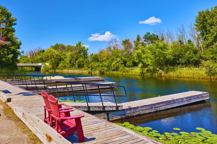 Two red muskoka chairs along the edge of a boardwalk by the dock slips on the Tay River
