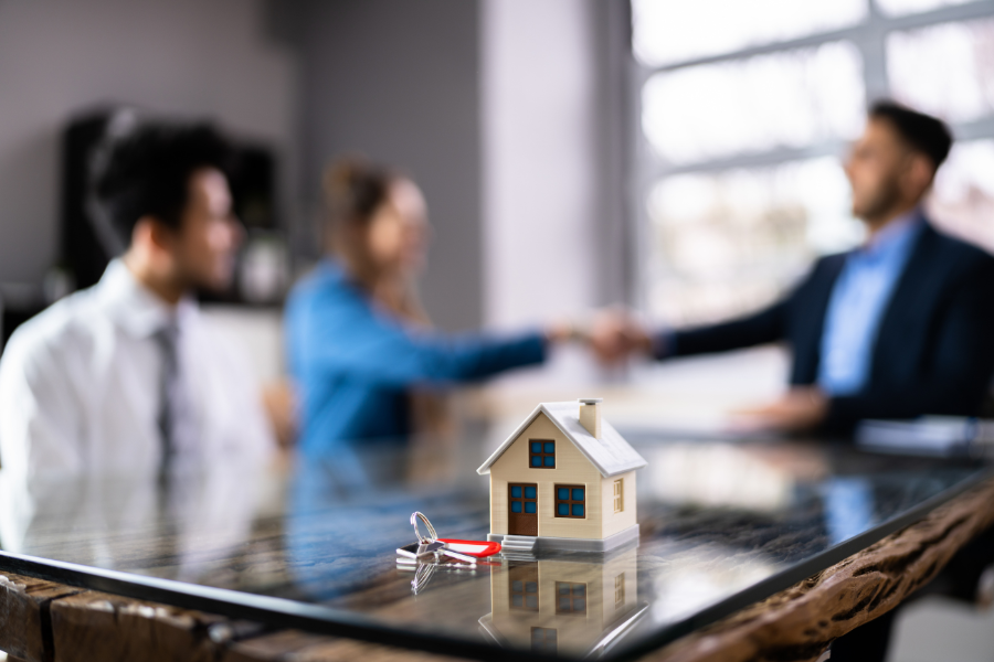 Small model of a house on a table next to a set of keys. A couple is in the background, with a woman shaking hands with another person