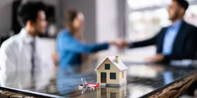 Small model of a house on a table next to a set of keys. A couple is in the background, with a woman shaking hands with another person