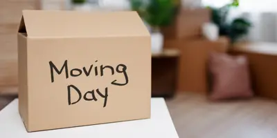Brown cardboard box with "Moving Day" written on it, sitting on top of a table. Various household items are in the background