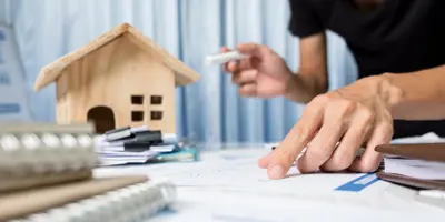Small model of a house next to a person holding a pen in one hand and pointing at a document on the table with the other. Several documents are in two stacks on the table in front of the house.